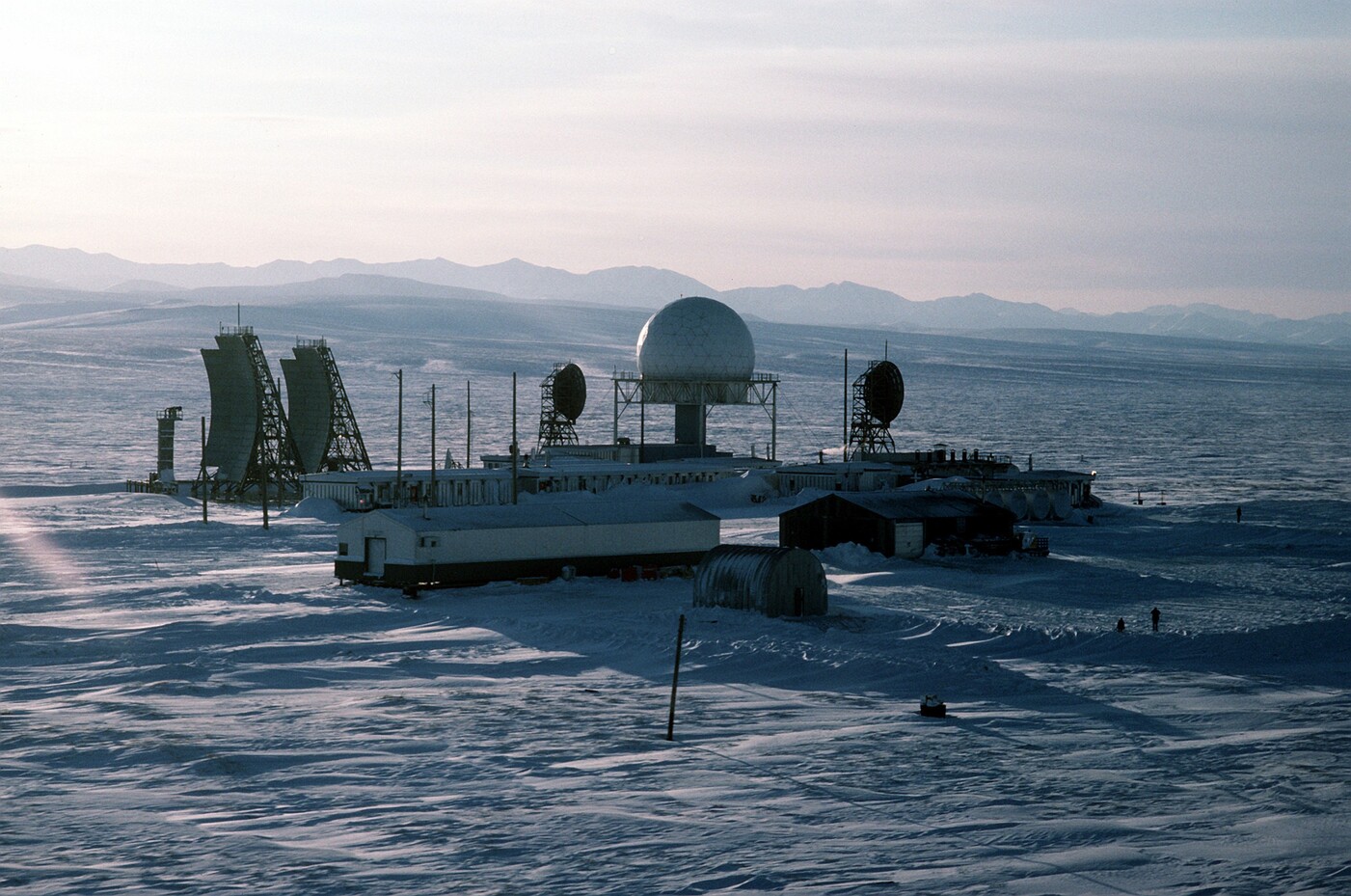 A DEW Line main station on the flat, snow-covered Alaskan Arctic coast in low sun: a white geodesic radome on a pedestal, two angled flat billboard antennas at left, round dish antennas, module-train buildings and a Quonset hut, hazy mountains behind, and two tiny figures for scale.