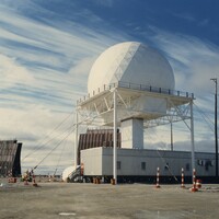 DEW Line radar station at Hall Beach, Northwest Territories, c. 1986 — a white geodesic radome on a tall open-steel tower above a corrugated module building, with a billboard tropospheric-scatter antenna alongside.