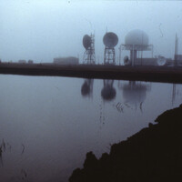The DEW Line Main station at Point Barrow, Alaska, reflected in a coastal lagoon in summer fog — tropospheric-scatter dish antennas on lattice towers, a geodesic radar radome, and low module buildings.