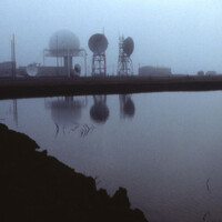 Point Barrow DEW Line station seen across a lagoon in fog — a geodesic radar radome and module buildings at left, tropospheric-scatter dish antennas on lattice towers at right.