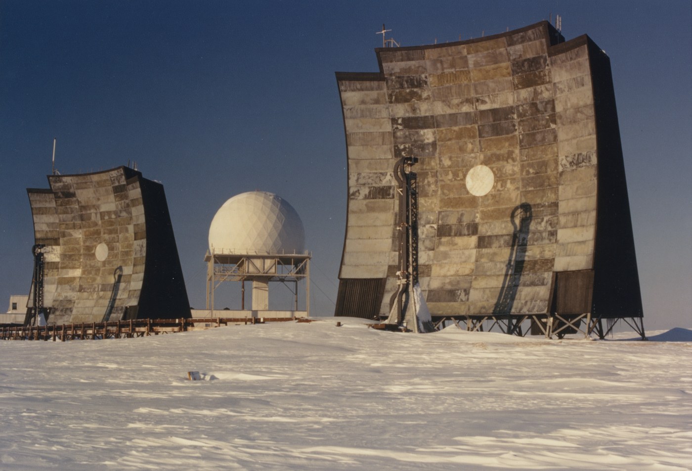 Two large flat billboard-style radar antennas flank a white geodesic radome at a DEW Line station in the Canadian Arctic, on snow under a deep blue sky.
