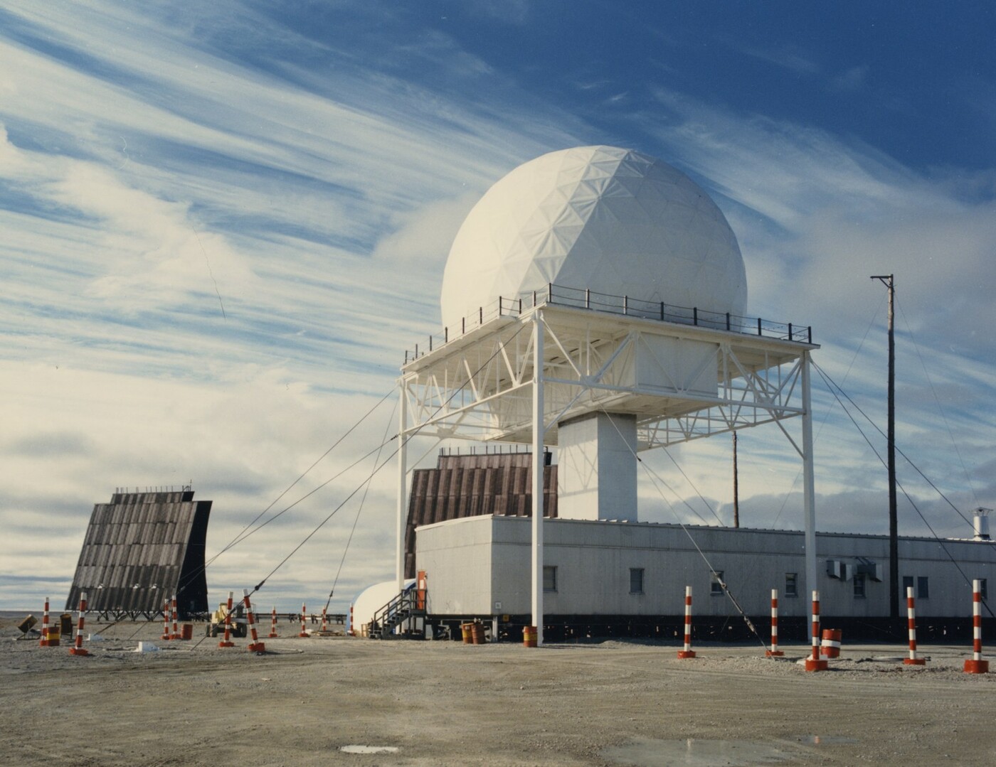 A DEW Line station under a blue Arctic sky streaked with high cirrus: a large white geodesic radome raised on an open white steel-frame tower above a long low building, a flat billboard search antenna angled at left, and red-and-white safety pylons on the bare gravel pad in the foreground.