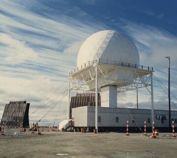 A DEW Line station under a blue Arctic sky streaked with high cirrus: a large white geodesic radome raised on an open white steel-frame tower above a long low building, a flat billboard search antenna angled at left, and red-and-white safety pylons on the bare gravel pad in the foreground.