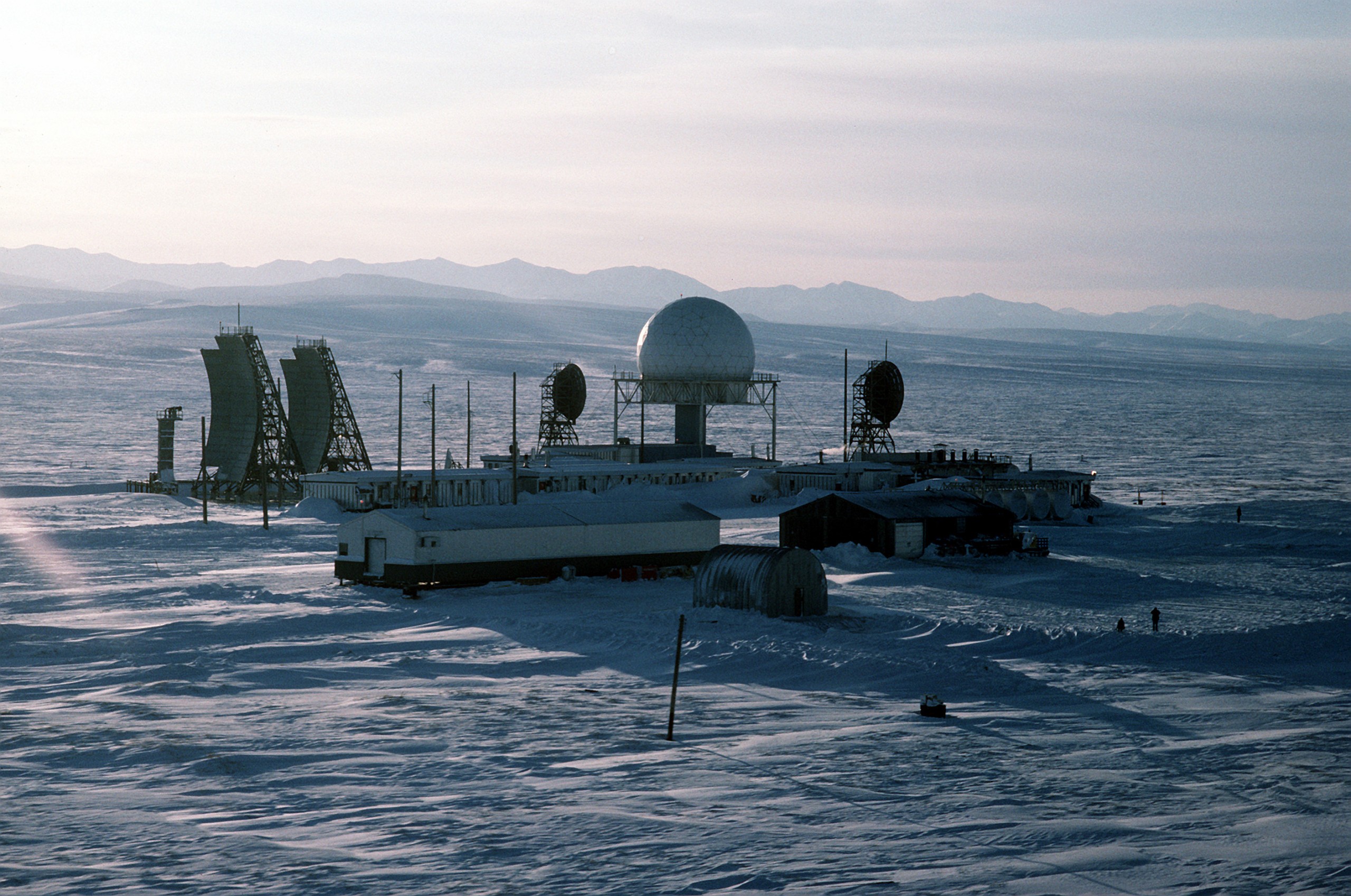 A DEW Line main station on the flat, snow-covered Alaskan Arctic coast in low sun: a white geodesic radome on a pedestal, two angled flat billboard antennas at left, round dish antennas, module-train buildings and a Quonset hut, hazy mountains behind, and two tiny figures for scale.
