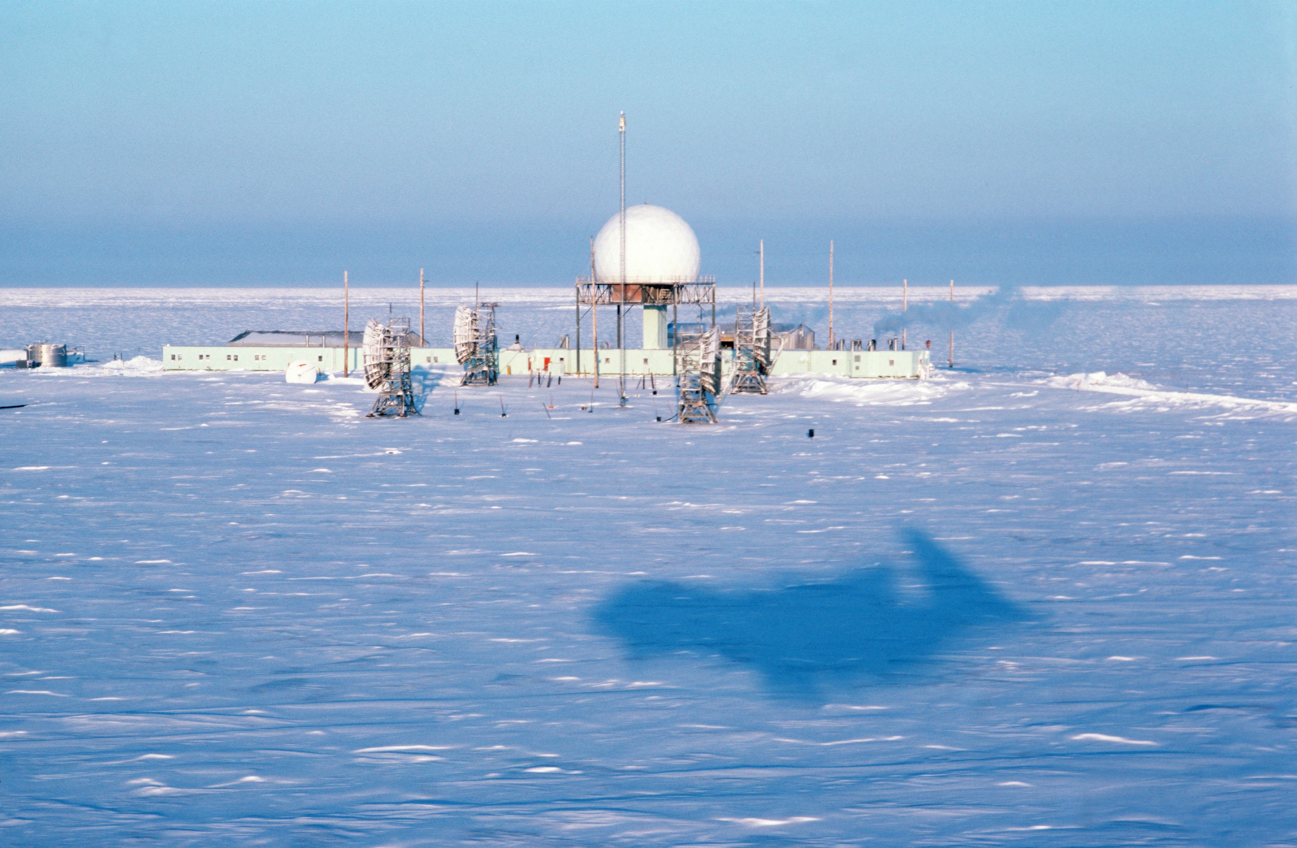 Aerial view of the POW-2 DEW Line radar station at Oliktok Point, Alaska, with its geodesic radome and module train buildings on the flat coastal tundra, the shadow of the photographing aircraft crossing the site.