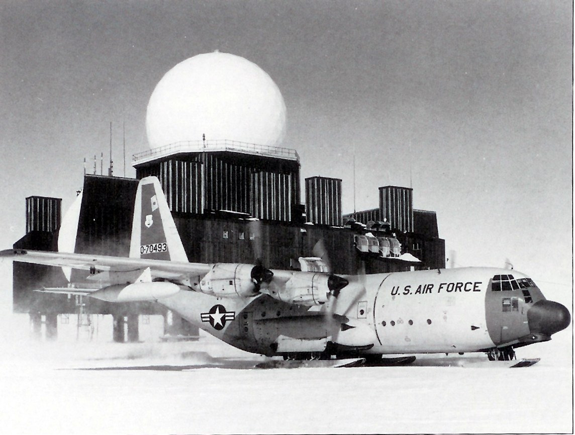 A ski-equipped U.S. Air Force C-130 Hercules transport parked on packed snow in front of a DEW Line station&rsquo;s module-train buildings and radome.