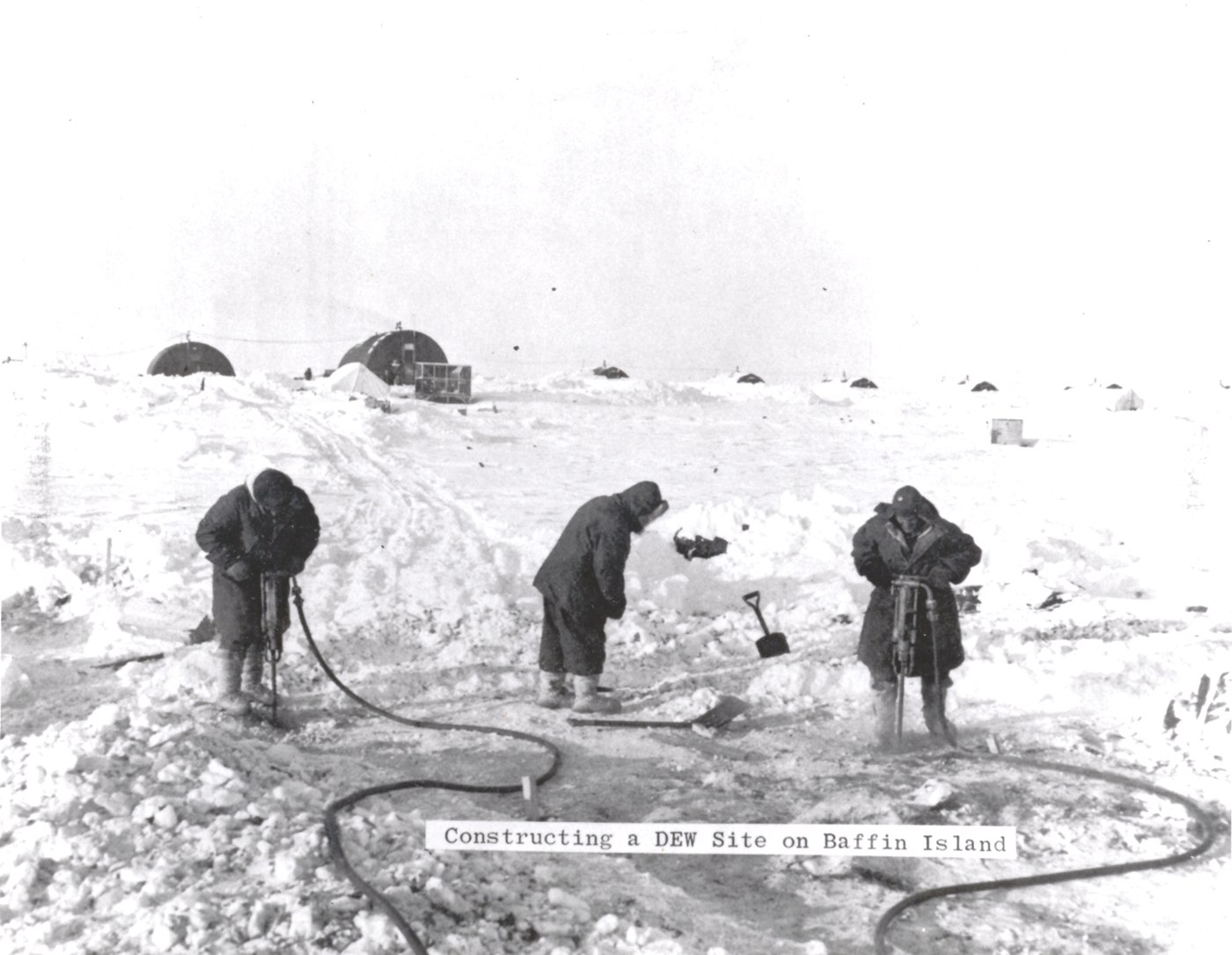 Three workers in heavy parkas break the frozen ground with a jackhammer and shovels at a DEW Line construction site on Baffin Island, prefabricated huts and snow behind them.