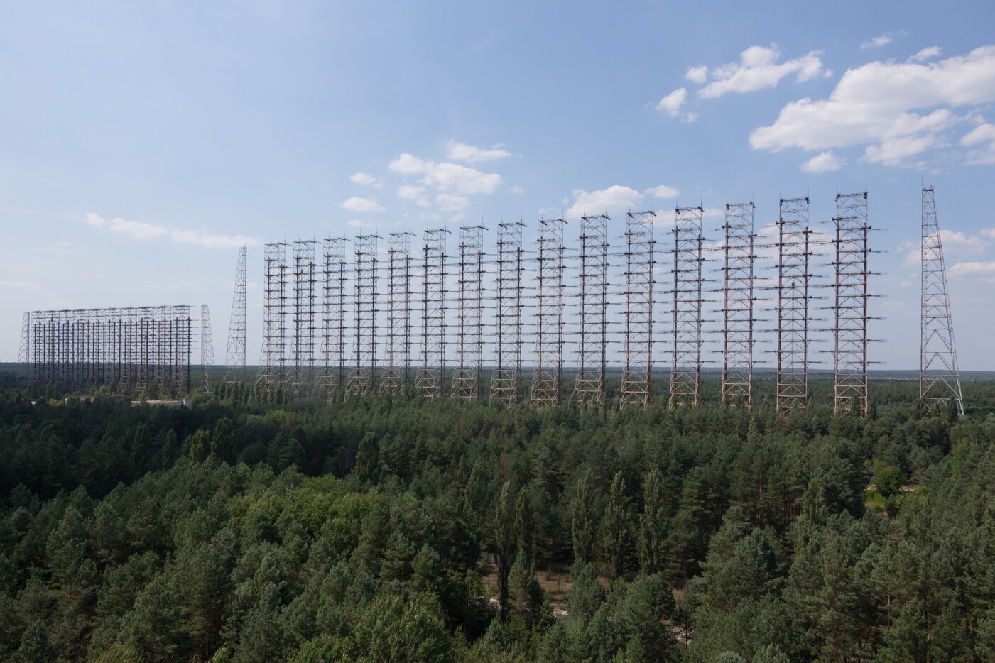 The abandoned Duga over-the-horizon radar near Chernobyl in 2014: a kilometers-long wall of rusting lattice towers strung with rows of cage antennas, rising above dense pine forest under a blue sky.