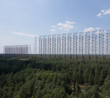 The abandoned Duga over-the-horizon radar near Chernobyl in 2014: a kilometers-long wall of rusting lattice towers strung with rows of cage antennas, rising above dense pine forest under a blue sky.
