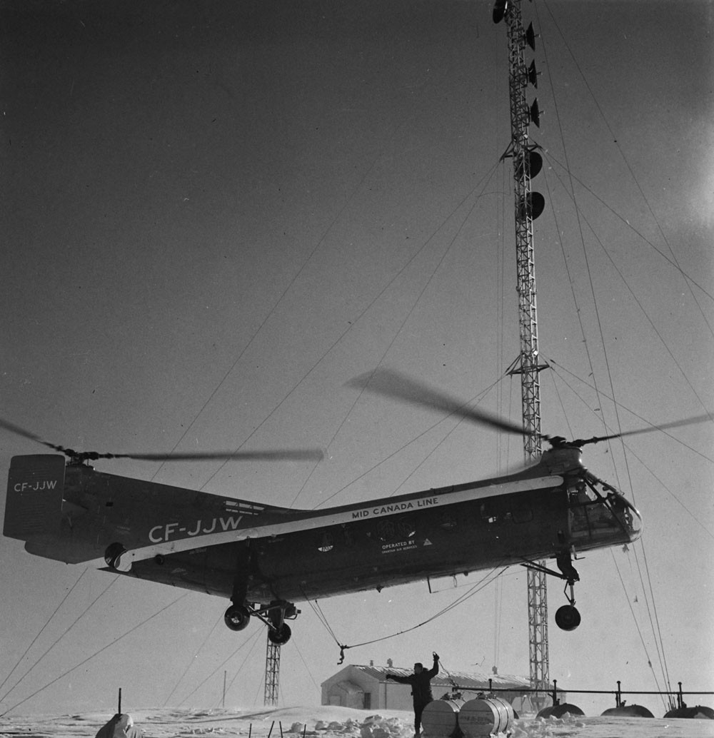 Black-and-white photo of a tandem-rotor Piasecki H-21 helicopter marked CF-JJW and Mid Canada Line hovering low over a snowy site, with a lattice tower carrying Doppler antennas and a worker guiding it in below.