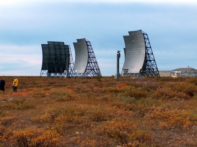 Muted-color view between the paired curved billboard antennas of a Mid-Canada Line sector station, weathered white operations buildings clustered between them on overgrown ground.