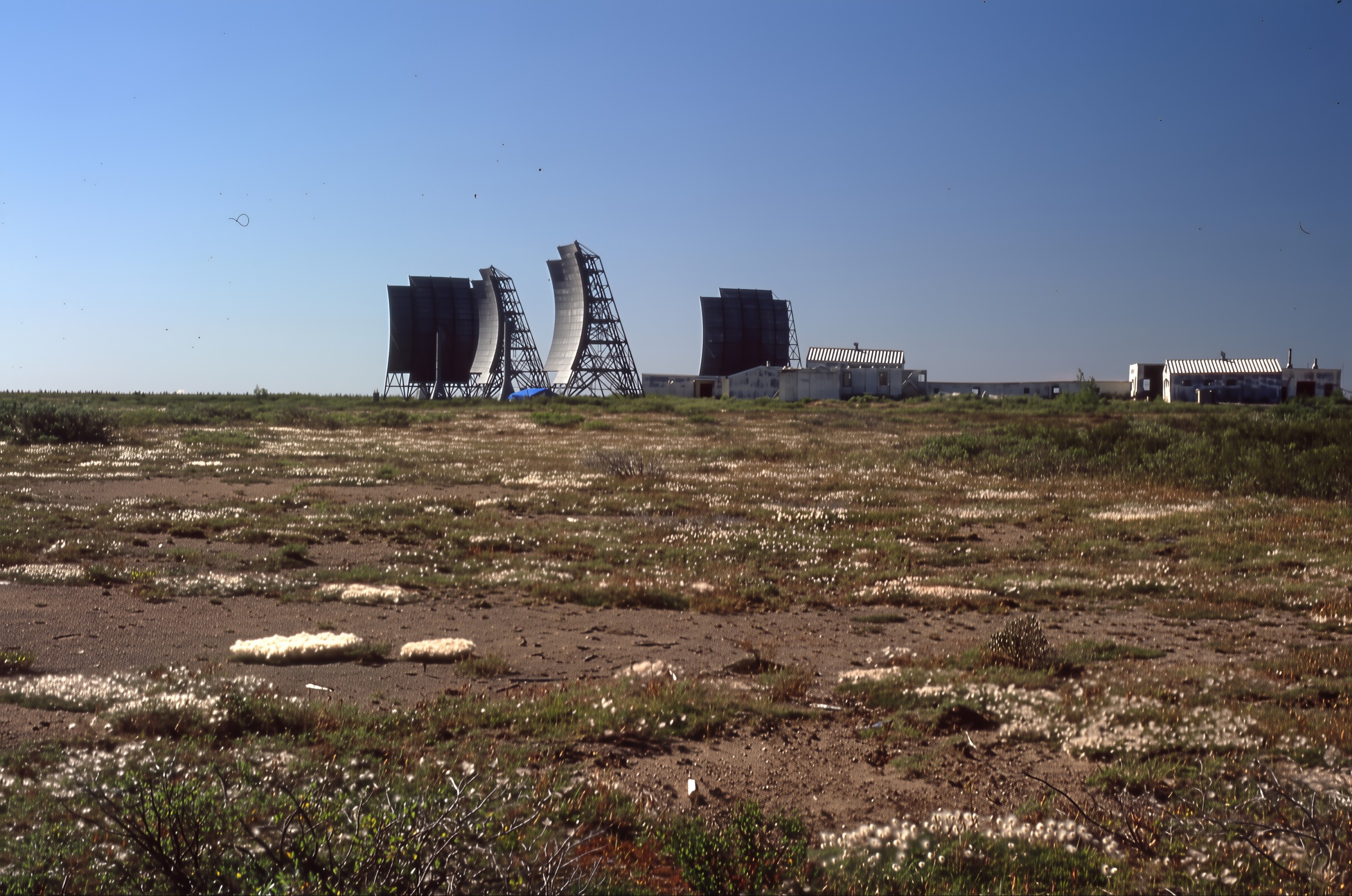 Abandoned Mid-Canada Line site on flat tundra: four curved troposcatter billboard antennas on steel lattice stands beside low white station buildings, seen across scrubby ground.