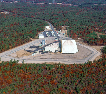 Aerial view of the PAVE PAWS radar at Cape Cod Air Force Station in 1986: a pale wedge-shaped building with two circular phased-array faces, ringed by a security fence in a clearing of autumn pine forest.